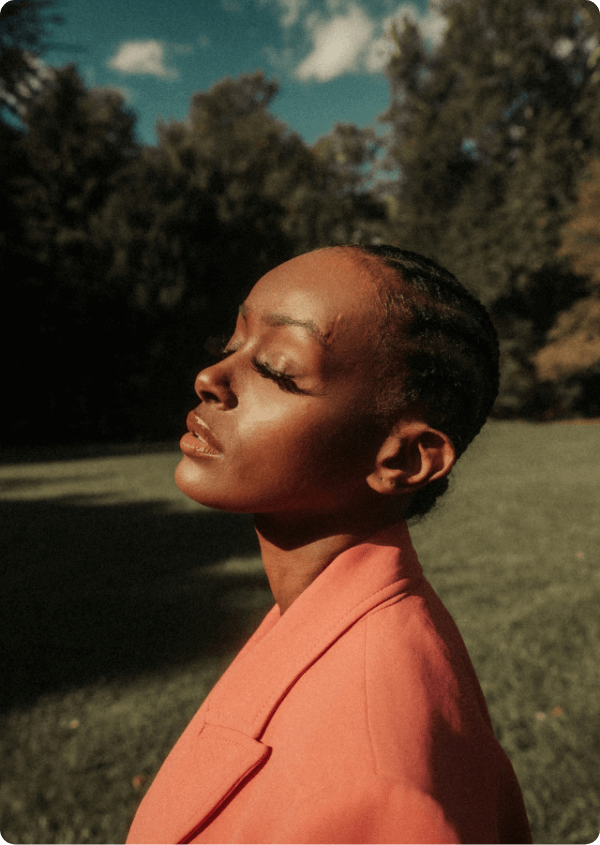 A woman looks to the sky with her eyes shut. She is in an orange blazer with greenery behind her
